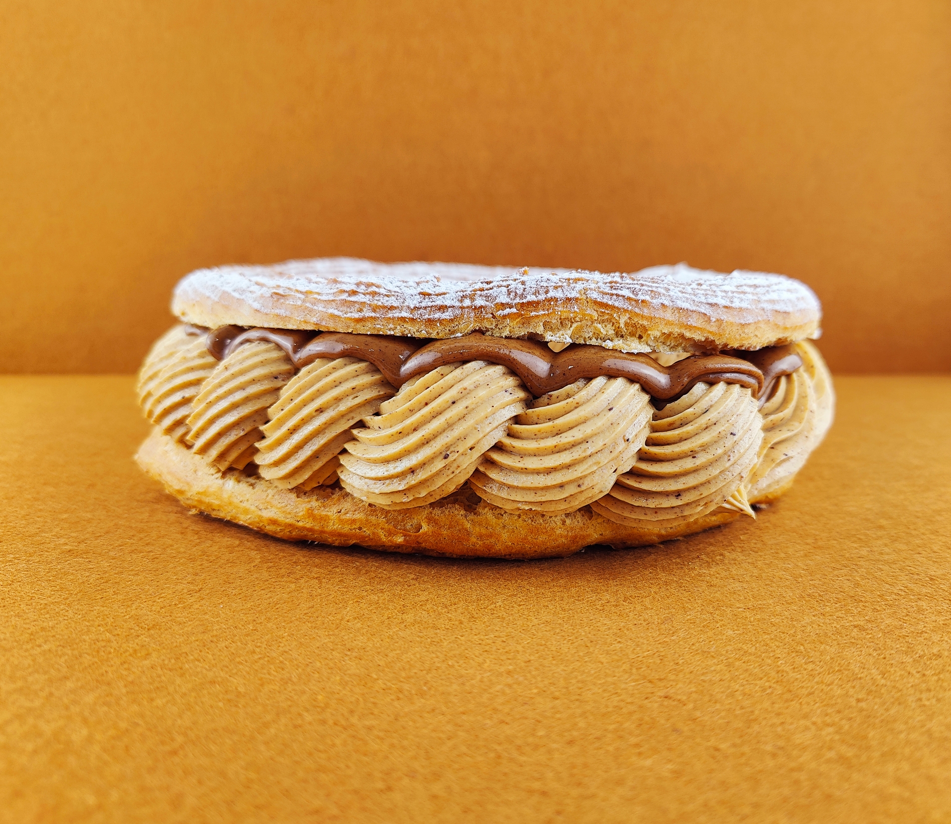 Tarta Paris-Brest vista de frente sobre fondo vacío de color marrón claro, mostrando el anillo de masa choux relleno de crema muselina de praliné con praliné encima, decorada con azúcar glas sobre la parte superior del anillo de masa choux.