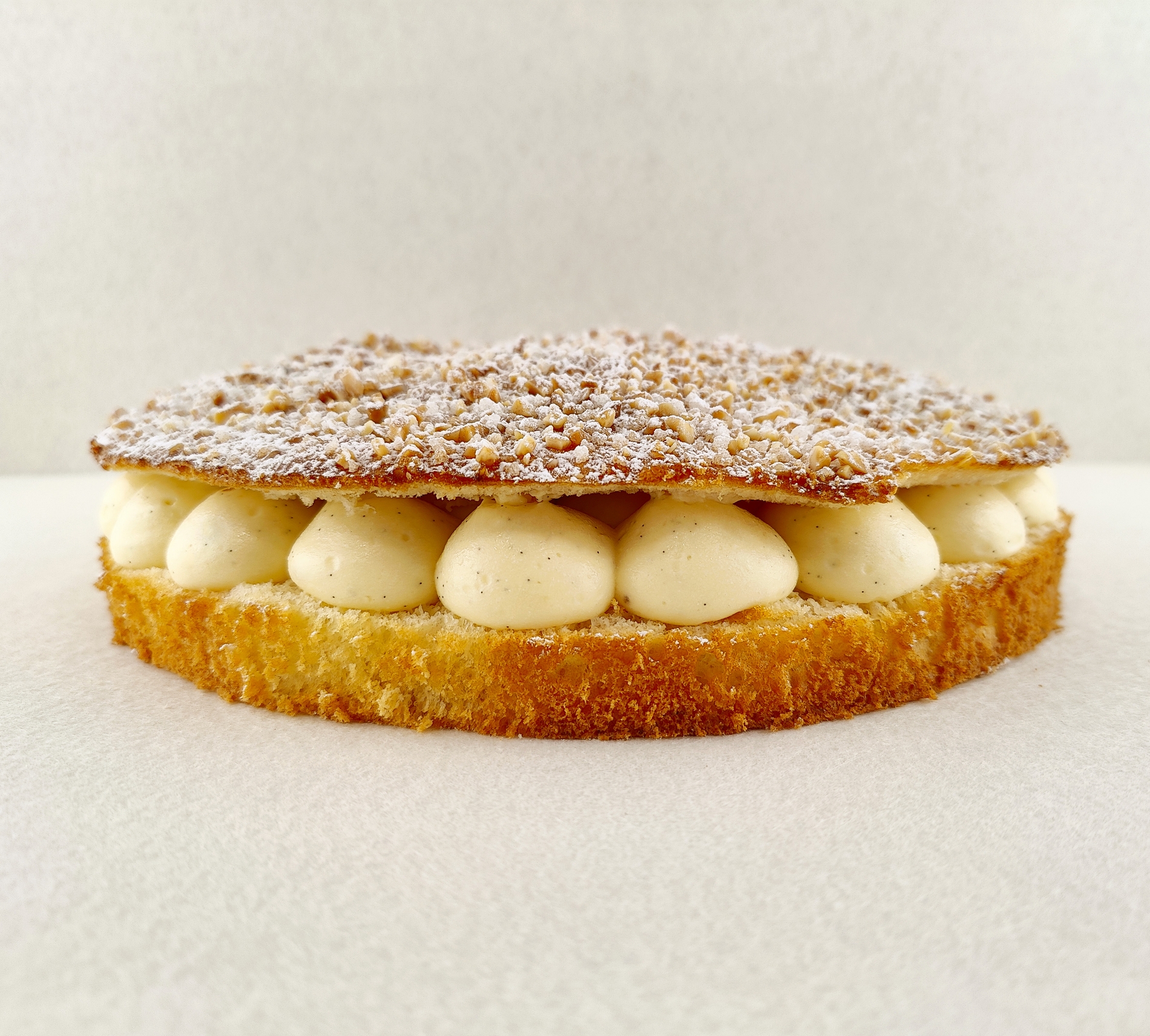 Tarta Tropézienne vista de frente, mostrando el brioche dorado relleno de crema diplomática a la vainilla, con la parte superior cubierta de azúcar perlado, almendra picada y azúcar glas, sobre fondo vacío y de color blanco roto.