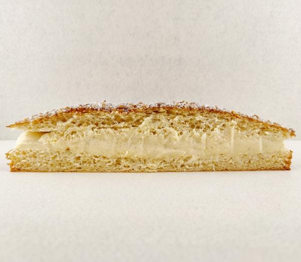 Tarta Tropézienne grande cortada a la mitad y fotografiada de frente, dejando ver el relleno de crema diplomática a la vainilla entre las capas de brioche, con la superficie espolvoreada de azúcar perlado, almendra picada y azúcar glas, sobre fondo vacío y de color blanco roto.