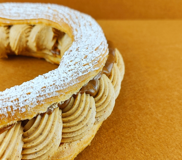 Primer plano de Tarta Paris-Brest sobre fondo vacío de color marrón claro, resaltando la textura de la crema muselina de praliné, el brillo del praliné y la cobertura de azúcar glas sobre el anillo de masa choux.