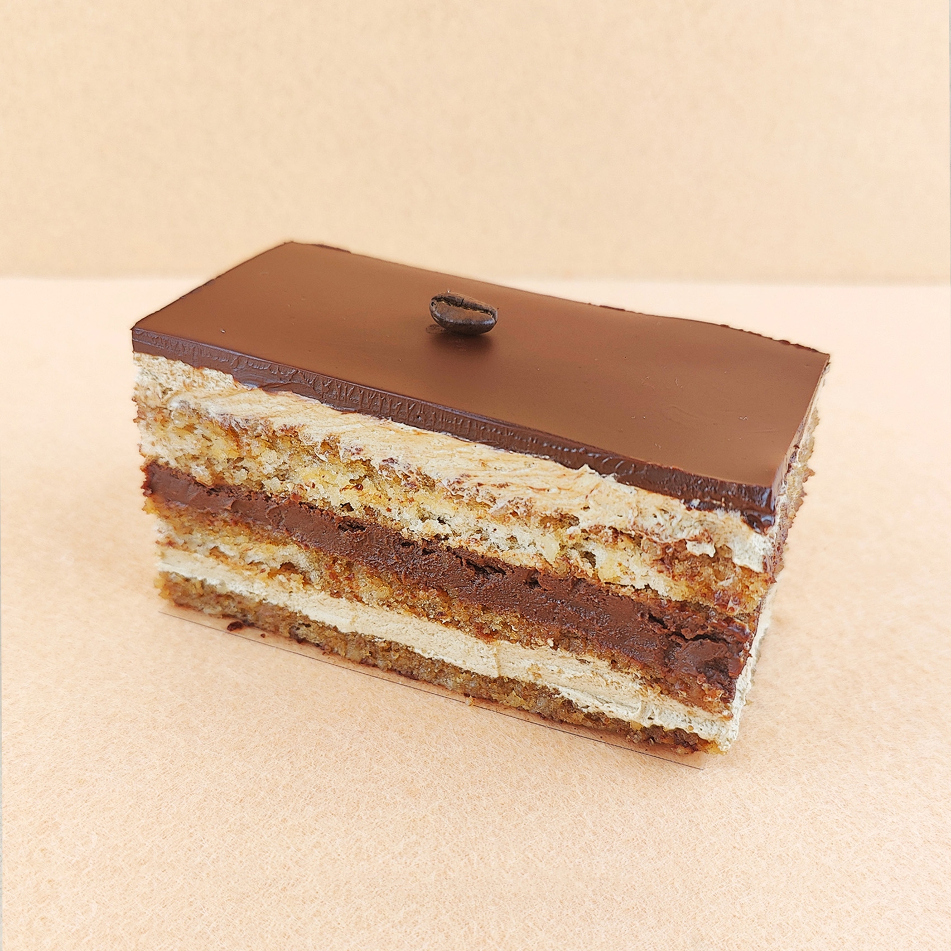 Porción individual rectangular de Tarta Ópera, fotografiada en ángulo de 45 grados con ligera inclinación, mostrando la cobertura de chocolate lisa y un grano de café decorativo en la parte superior.