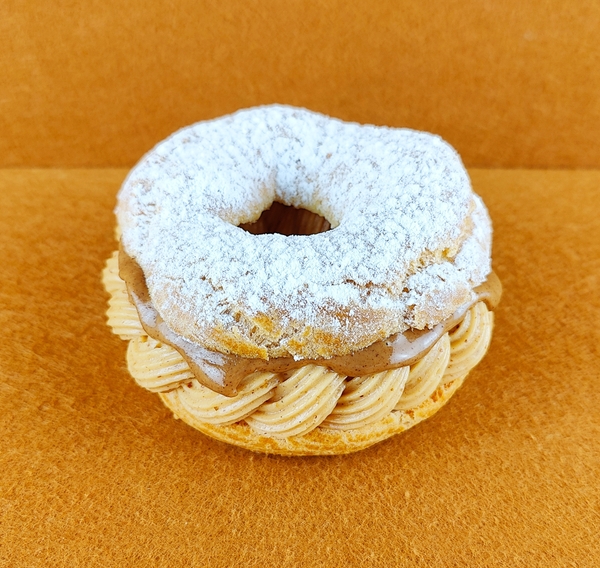 Tarta Paris-Brest individual fotografiada en ángulo de 45 grados sobre fondo vacío de color marrón claro, con anillo de masa choux relleno de crema muselina de praliné y praliné por encima, decorada con azúcar glas sobre la parte superior del anillo de masa choux.