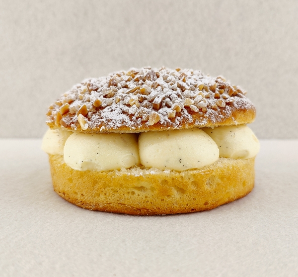 Tarta Tropézienne pequeña individual vista de frente, mostrando el brioche dorado relleno de crema diplomática a la vainilla, con la parte superior cubierta de azúcar perlado, almendra picada y azúcar glas, sobre fondo vacío y de color blanco roto.