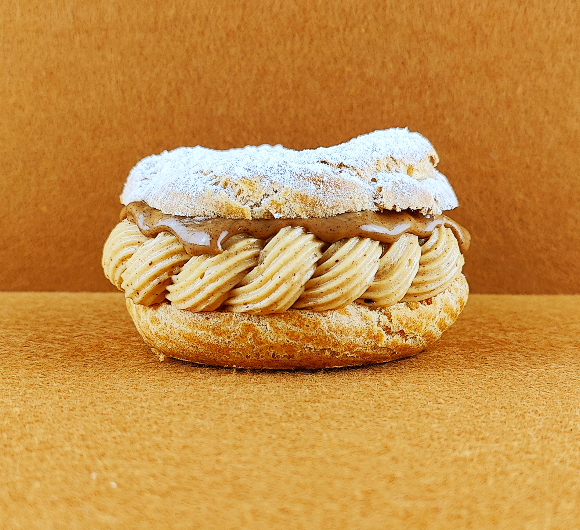 Tarta Paris-Brest individual vista de frente sobre fondo vacío de color marrón claro, mostrando el anillo de masa choux relleno de crema muselina de praliné con praliné encima, decorada con azúcar glas sobre la parte superior del anillo de masa choux.